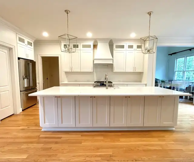White kitchen with a long shaker island, a white range hood, brass lantern pendants and oak floors