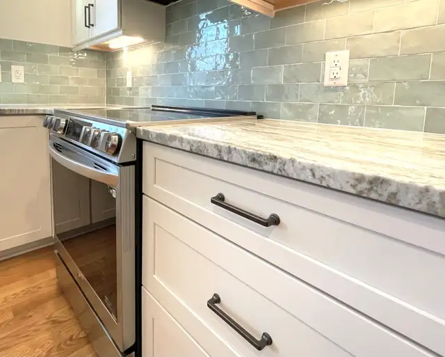 Close-up of a quartzite countertop edge over white shaker drawers with bronze pulls and a green glazed tile backsplash